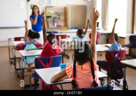 Vista posteriore degli studenti che alzano le mani mentre si siedono sulla scrivania in classe a scuola Foto Stock