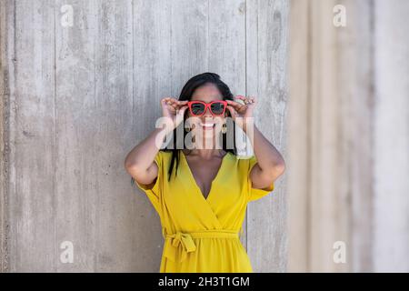 Vista frontale vicino a una colonna di una giovane donna latina in abito giallo e occhiali da sole che le tengono con entrambe le mani e guardando la macchina fotografica contro un muro a d Foto Stock