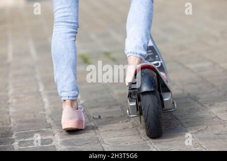 Primo piano di piedi femminili di una ragazza che indossa sneakers rosa e un paio di jeans su uno scooter elettrico sullo sfondo delle strade della città Foto Stock