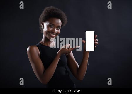 Sorridente donna nera che punta con il dito su un modello vuoto cellulare schermo bianco Foto Stock