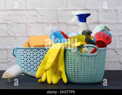 Spugne, spazzole di plastica e bottiglie di detersivi su un tavolo di legno blu. Articoli per la pulizia della casa su sfondo bianco della parete in mattoni Foto Stock