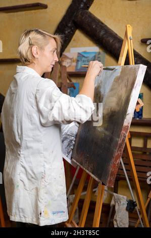 Donna felice dipinge su tela usando pennello sottile. La tela si trova sul cavalletto. L'artista disegna al cavalletto. Vista laterale della pittura femminile p Foto Stock