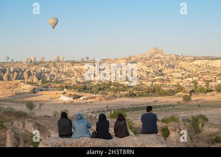 GOREME, TURCHIA - 5 AGOSTO 2021: La gente guarda al mattino seduto sulle rocce, come mongolfiere volare i turisti sulla valle della Cappadocia con unità Centrale Abitacolo Foto Stock