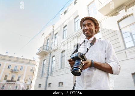 Uomo nero con fotocamera digitale in piedi nella città europea. Giovane ragazzo sorridente indossa un cappello di paglia, una camicia bianca e un orologio da polso. Concetto di viaggio. Idea di Foto Stock