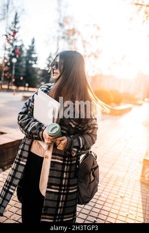 Bella giovane studentessa con caffè, libri e uno zaino che trascorre del tempo al parco, musica con cuffie Foto Stock