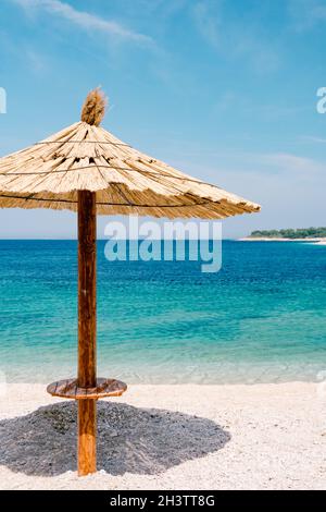 Un ombrello spiaggia con tetto di paglia contro il cielo blu e l'acqua azzurra su una spiaggia di sabbia in Croazia, nella città di Primosten. Foto Stock