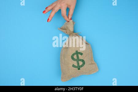 La mano tiene un sacchetto di tela pieno di soldi con un'icona del dollaro degli Stati Uniti su uno sfondo blu Foto Stock