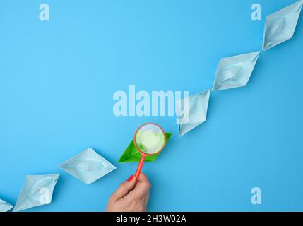 La mano femmina tiene una lente d'ingrandimento rossa su una fila di barche di carta su sfondo blu Foto Stock