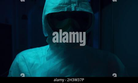 Dettagli della ricerca horror. Primo piano di persona sconosciuta in tuta protettiva e maschera, spaventosamente in piedi nella sala con luce lampeggiante. Foto Stock
