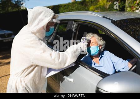 Lavoratore medico che indossa tuta ppe prendendo la temperatura di donna caucasica anziana seduto in auto Foto Stock