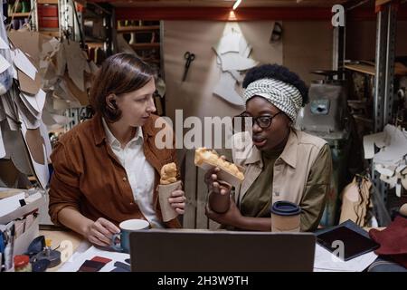 Due cassettiere seduti al tavolo e che si comunicano a pranzo con panini e bevande a base di caffè in officina Foto Stock