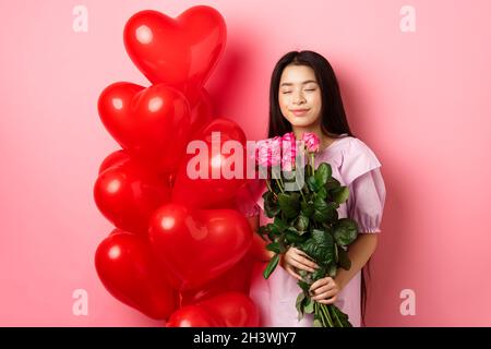 Concetto di giorno di San Valentino. Romantica ragazza asiatica teen sognando di amore o data, occhi vicini e sorriso, tenendo fiori da amante, rec Foto Stock