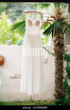 L'abito da sposa con un corsone trasparente in pizzo e un corpetto beige su un appendiabiti vicino a una recinzione in pietra bianca e un decoro Foto Stock