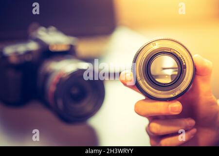 Il fotografo tiene in mano un obiettivo fotografico, una fotocamera e un computer portatile in uno sfondo sfocato Foto Stock