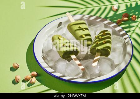 Pistacchio surgelato fatto in casa in ciotola di ghiaccio su sfondo verde con ombra di foglia di palma in luce dura. Aggiornamento della pop-sicle, Foto Stock