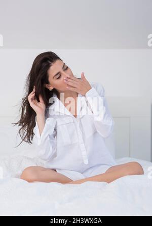 Giovane donna con lunghi capelli neri. È seduta sul e indossa una camicia bianca. Ha una mano davanti alla sua bocca e sbadigia. Foto Stock