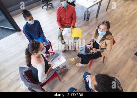 Un gruppo eterogeneo di colleghi creativi che indossano maschere facciali che parlano durante la riunione Foto Stock