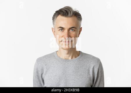 Primo piano di uomo di mezza età sicuro e bello con acconciatura corta grigia, sorridente e deciso, in piedi su bianco b Foto Stock