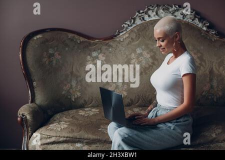 Giovane donna millenaria con ritratto corto biondo con computer portatile sul divano vintage. Foto Stock