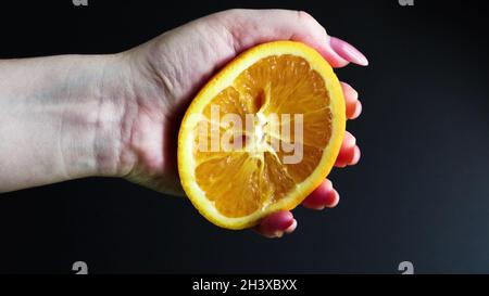 Una mano femminile spreme metà di un arancio fresco e succoso. Succo d'arancia di agrumi gocciolando dalla polpa d'arancia su sfondo nero. Th Foto Stock