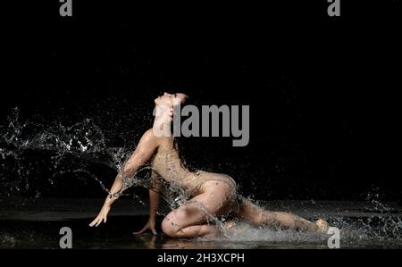 Giovane donna con capelli neri lunghi giace a terra sotto le gocce di pioggia su sfondo nero. Donna vestita con bodysuit beige Foto Stock