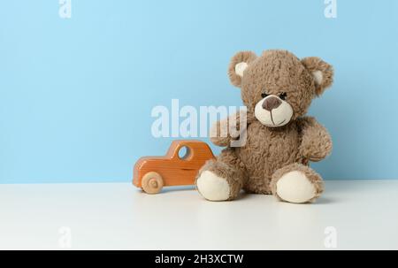 Il grazioso orsacchiotto marrone si siede su un tavolo bianco e un'auto giocattolo in legno per bambini, sfondo blu Foto Stock