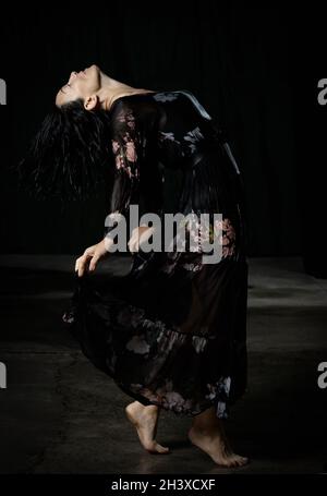 Giovane donna con capelli neri danze in un abito nero a chiffon lungo bagnato su sfondo scuro Foto Stock