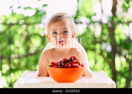 Il bambino con una faccia sporca si siede ad un tavolo davanti ad un piatto di frutta Foto Stock