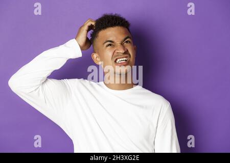 Primo piano di un uomo afro-americano travagliato che pensa, graffia la testa e guarda in alto confuso, in piedi in maglia bianca sopra la borsetta Foto Stock