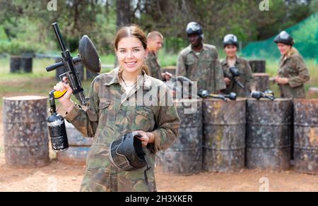 Giovane giocatore di paintball femminile in camouflage in piedi sul campo di battaglia con la pistola prima di giocare Foto Stock