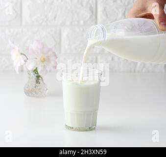 Processo di versare il latte fresco da una bottiglia di plastica in una tazza di vetro, tavola bianca Foto Stock