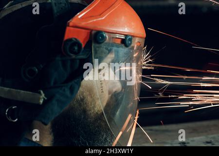 Uomo in maschera protettiva trasparente lavora su metallo con sega circolare. Scintille e faccia. Foto Stock