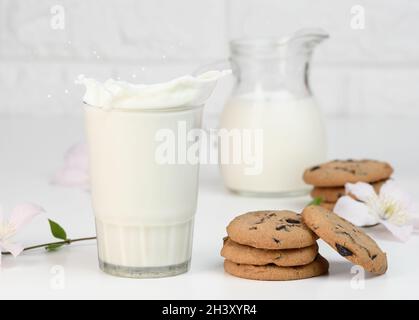 Una tazza di vetro piena di latte fresco con spruzzi si trova su un tavolo bianco, colazione sana Foto Stock