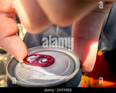 Primo piano dell'uomo che tiene e apre la linguetta tirata della lattina di birra o di soda. Mani aperte una bevanda in alluminio metallo lattine cola con luce naturale del sole a te Foto Stock