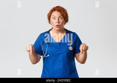 Medicina, sanità e coronavirus concetto. Impressionato e eccitato rosso medio-anziano medico di donna in scrub, puntando il dito Foto Stock
