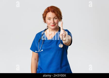 Medicina, sanità e coronavirus concetto. Sorridente femmina medico di mezza età, medico in scrub scuotendo il dito in WARN Foto Stock
