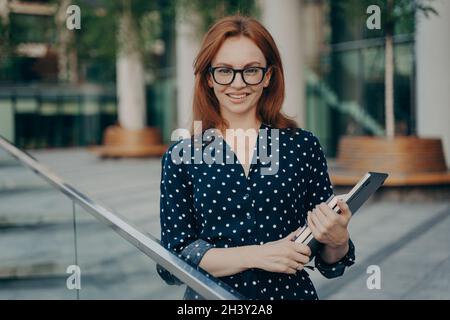 Prospera donna d'affari pone all'aperto in elegante vestito trasporta gli articoli necessari per il lavoro Foto Stock