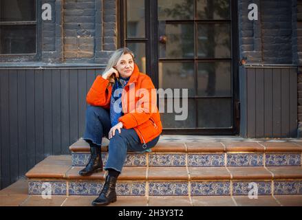 Felice sorridente donna di mezza età in giacca arancione giù seduta su scale di cemento all'aperto Foto Stock