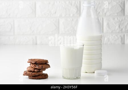 Una tazza di vetro piena di latte e una bottiglia di plastica trasparente con latte, accanto a una pila di biscotti rotondi con scaglie di cioccolato su un tavolo bianco Foto Stock