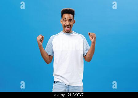 Sostenente e allegro giovane sportivo afroamericano in t-shirt alla moda che alza i pugni in vittoria e trionfo gesto urlare Foto Stock