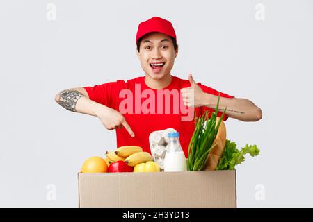 Shopping in linea, consegna del cibo e concetto dei depositi del Internet. Sorridendo il corriere asiatico in t-shirt rossa e cappellino, mostra il pollice-up di nuovo Foto Stock