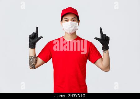 Fidato giovane uomo asiatico in testa rossa e t-shirt, indossare maschera medica protettiva e guanti, puntando le dita verso l'alto, mostra Foto Stock