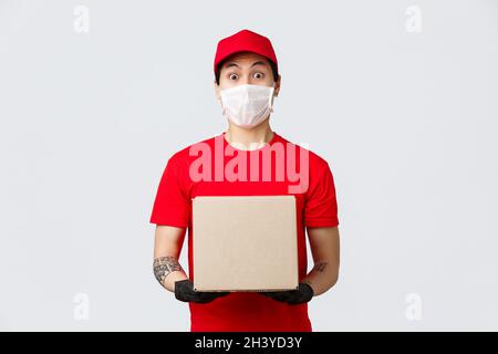 Sorpresi corriere in rosso uniforme cappuccio e t-shirt, custodia con ordine cliente, indossare maschera medica e guanti protettivi per Foto Stock