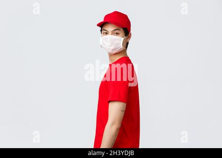 Ritratto del ragazzo di consegna asiatico in berretto rosso e t-shirt, indossare maschera medica protettiva, girare faccia a macchina fotografica. Corriere in uniforme del Foto Stock