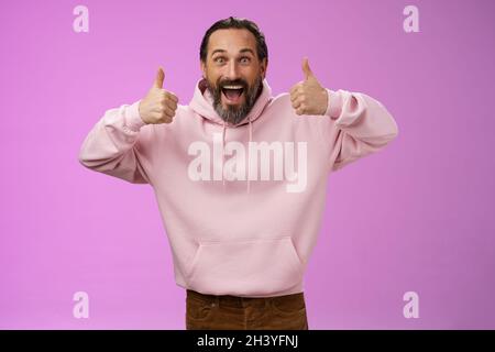 Eccitata energica carismatica europea matura uomo bearded in rosa felpa con cappuccio mostra pollici verso l'alto gesto sorridente acconsento fornisce Foto Stock