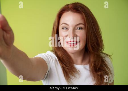 Primo piano allegro entusiasta rossa affascinante mezza età donna estando braccio verso fotocamera che prende selfie smartphone sorridendo ampio Foto Stock