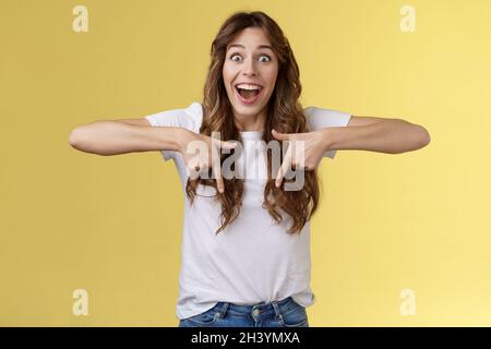 Eccitato felice allegro impressionato cute brunette ragazza lungo taglio di capelli sorridente ampiamente sorpreso reagire felice indicando verso il basso Share ecc Foto Stock