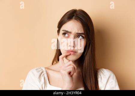 Ritratto di ragazza premurosa accigliata e guardando da parte pensiva, pensando o facendo scelta, in piedi su sfondo beige Foto Stock