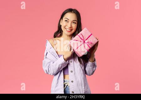 Curioso che cosa è dentro. Ritratto di carino ragazza asiatica sciocca scuotendo scatola con regalo per indovinare che cosa è esso, sorridendo divertito, appoggiandosi al fotoricettore Foto Stock