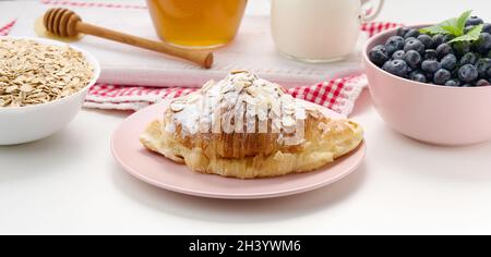 Croissant al forno cosparso di zucchero in polvere, mirtilli e farinata d'avena in un piatto di ceramica su un tavolo bianco Foto Stock